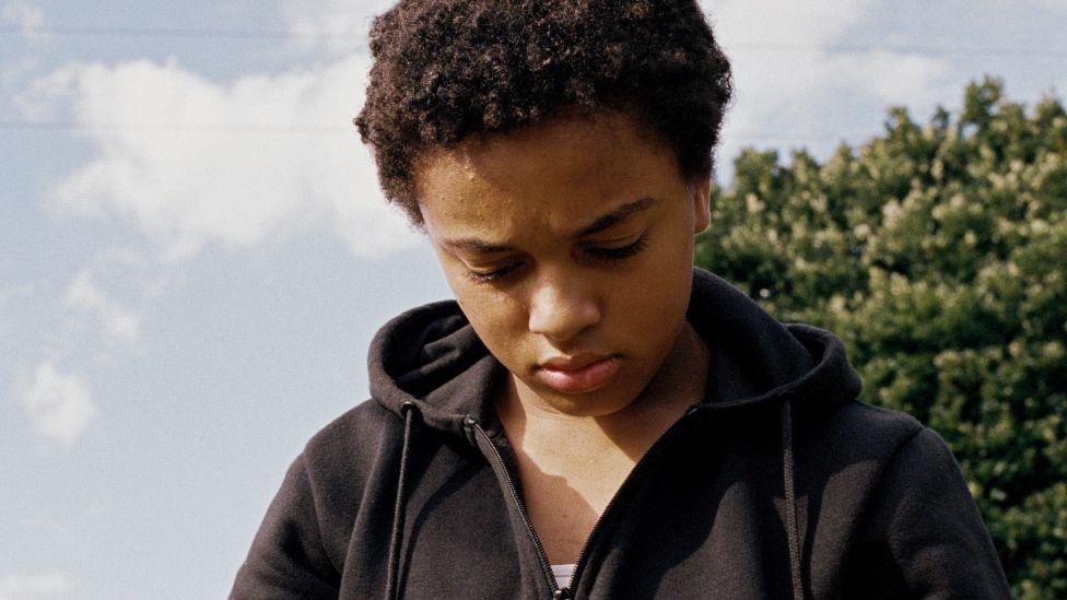 Nykiya Adams as Bailey in Bird, pictured in a black hoodie and looking at the ground