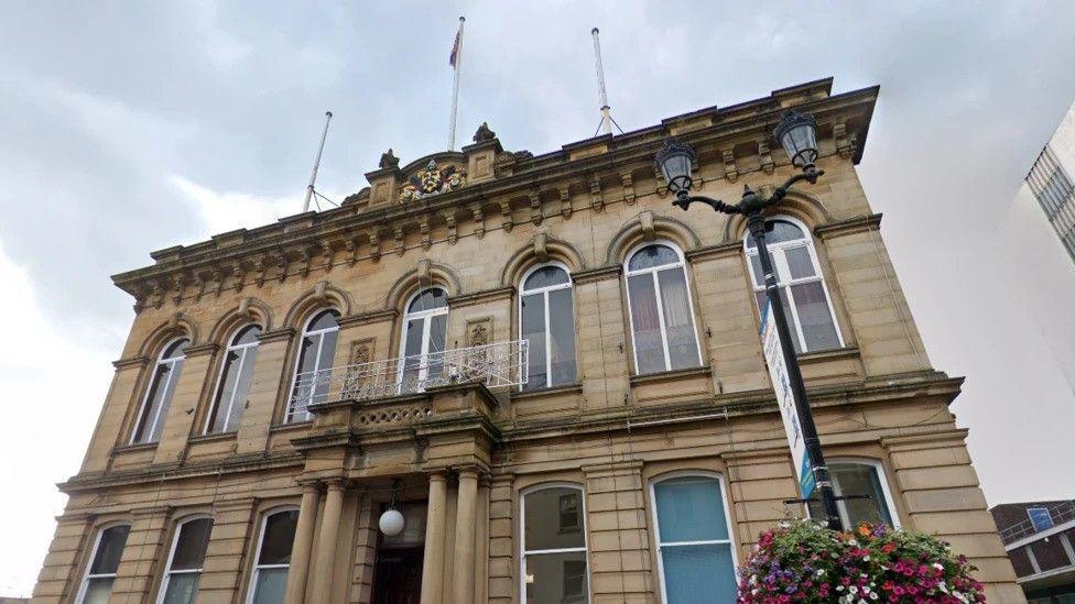 A town hall, pictured on cloudy day. A hanging basket of flowers sits beneath a sign outside.