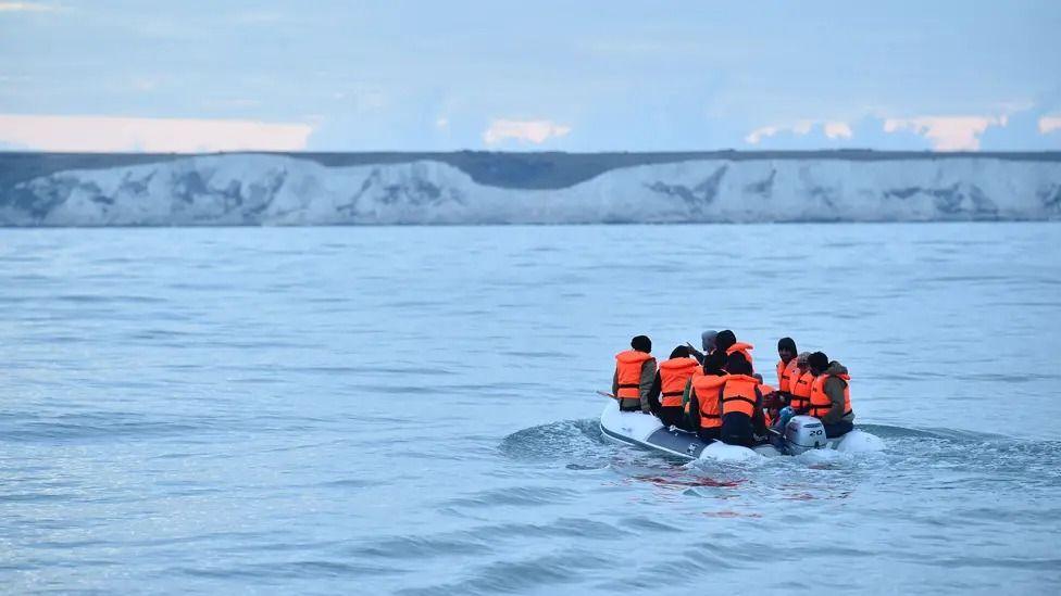 A group of about nine or 10 people in an inflatable boat, wearing life jackets. In the background are the white cliffs of the south coast of England