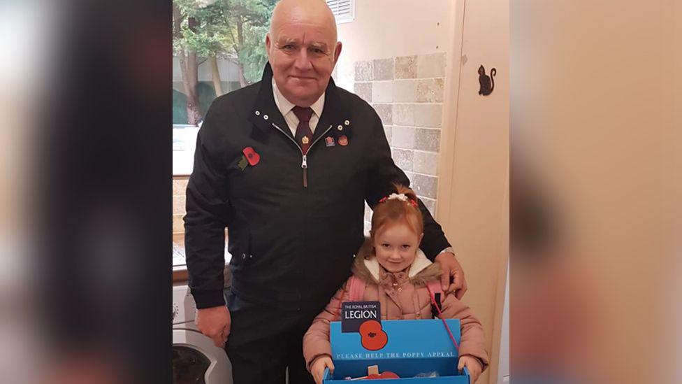 A young Olivia and her grandfather Terry Clarkson posing. Olivia is  comes up to her grandfather's elbow and holding a large blue box of poppies.