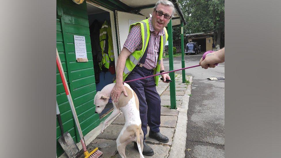 Brian Johnson - wearing dark blue trousers and a red and white checked shirt with a hi-vis sleeveless jacket over the top - with a beige and cream coloured dog leaping up his leg