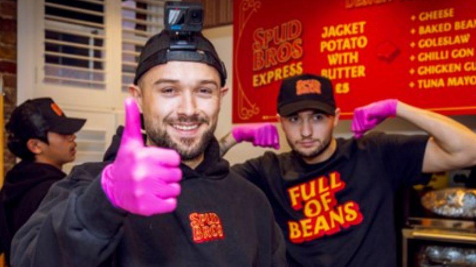 Jacob (left) and Harley Nelson in the kitchen in front of a red menu. Both are wearing black baseball hats and tops. Jacob is doing a thumbs up gesture while wearing a pink latex glove and Harley is in the background flexing his muscles