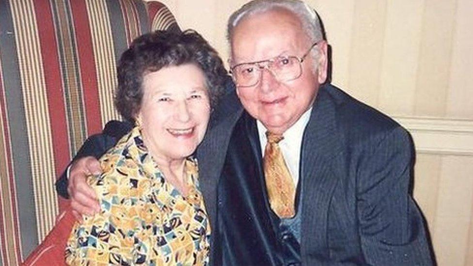 A brown-haired woman wearing a yellow-patterned dress sitting sitting next to a grey haired man, who is wearing a blue jacket and blue waistcoat and yellow tie. The man has his right arm around the woman's shoulders.