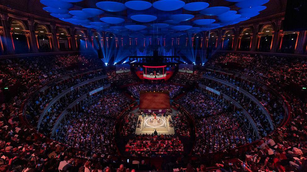 A packed arena with tiered seating surrounds a brightly lit boxing ring at the center. Blue circular panels hang from the ceiling, and dramatic lighting highlights the venue’s grand architecture.