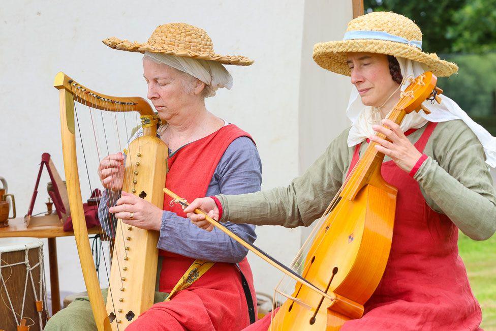Two women in historic costume play old stringed instruments one that looks like a harp and the other more a cello-type instrument