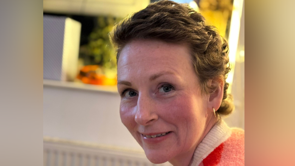 Claire is in a home and is looking over her left shoulder at the camera while smiling slightly. She has short brown wavy hair and is wearing gold hoop earrings. She has a coral coloured jumper with with a white collar and behind her is an out-of-focus kitchen.