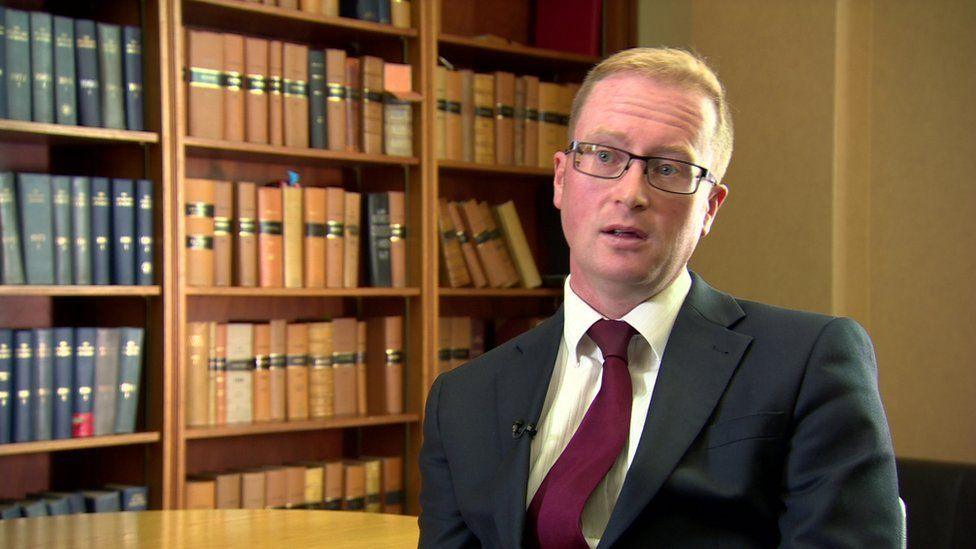 Joe McCrisken, who has red hair and glasses and is wearing a suit, sits in front of a library of books