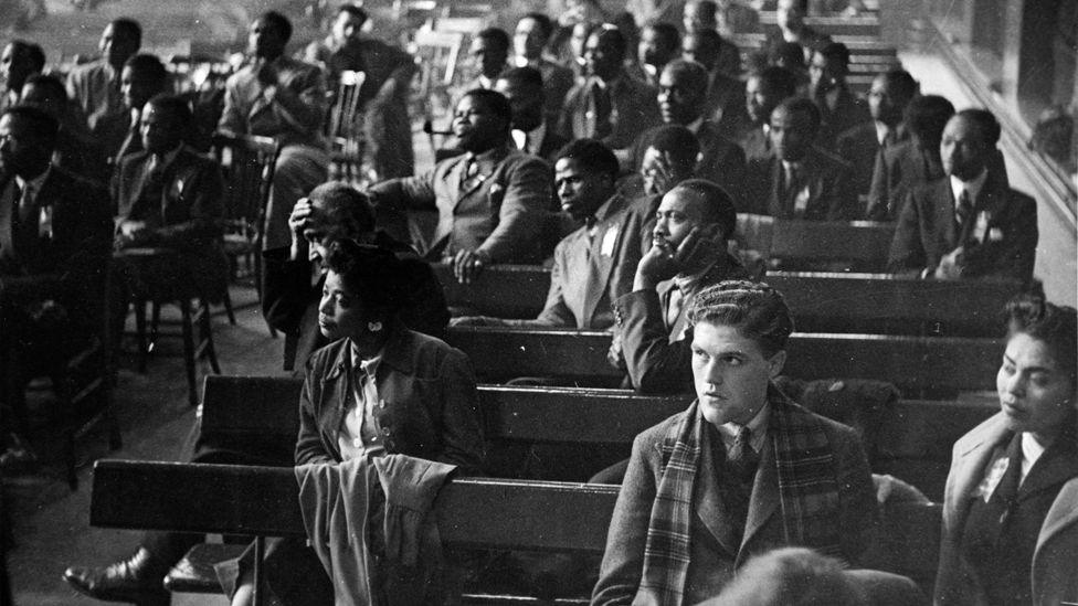 The audience of black and white attendees, wearing smart clothes and sitting on wooden benches, listening to speakers at the Fifth Pan-African Congress, held at Chorlton-upon-Medlock Town Hall in Manchester