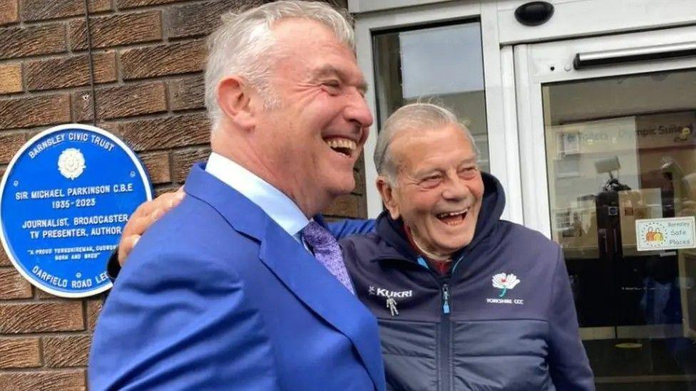 Two men, one seemingly in his fifties and the other in his eighties, each with one arm around the other's shoulder, laughing at something outside of shot. The younger man wears a blue suit, and the elder a navy cricket zip-up jacket.