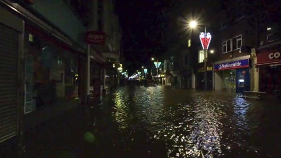An image of a high street at night that has flooded. There are shops but the street is covered in water. There are also street lights that are on. 