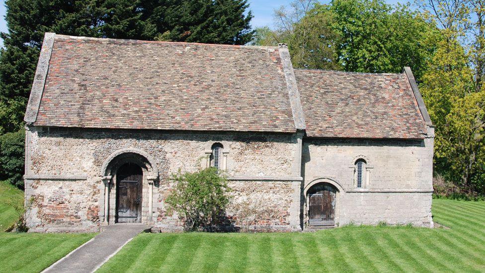 The Cambridge Leper Chapel, a stone-built medieval buildings with two arched doorways and two narrow arched windows and a tiled roof, with trees in leaf behind and mowed grass in front