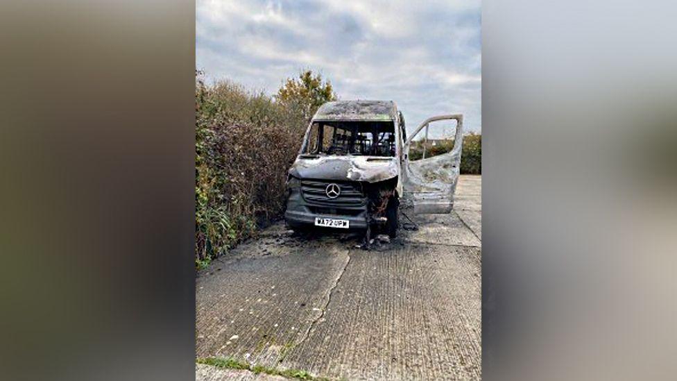 White Mercedes minibus destroyed by fire.  The inside of the bus is completely burned out.  The passenger door is open and there is some damage to the frontage.  It is parked on concrete with foliage in the background.