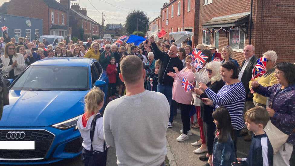 Scores of people in the street waving bunting and Union Jack flags as Maise arrives home in a blue car