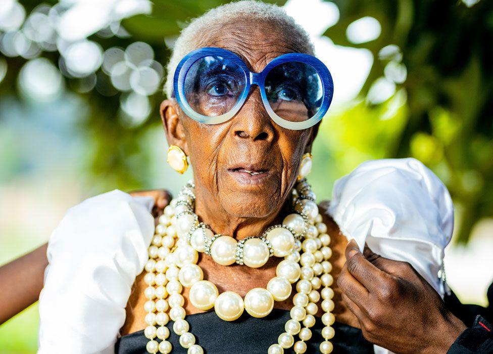 A  hand can be seen adjusting one of Margret Chola's puffed white sleeves. She is wearing big blue-framed sunglasses and four strings of pearls of various sizes