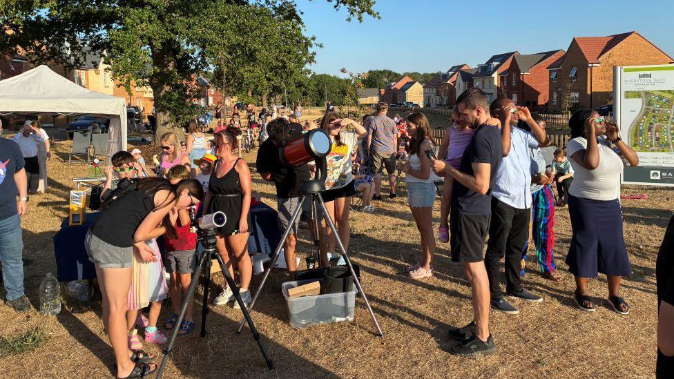A crowd of people of all ages are gathered together and looking at telescopes in a housing estate, on a green.