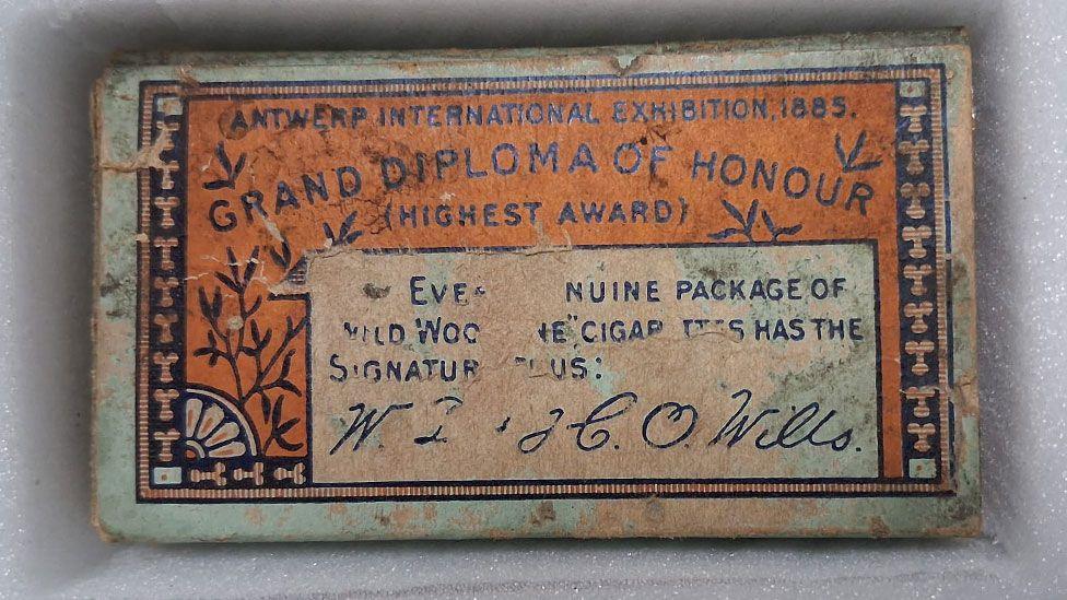 A rather battered cigarette box. It is rectangular with an orange and black pattern and says in capital letters Antwerp International Exhibition 1885 grand diploma of honour (highest award) along the top. Beneath in a cream rectangle are other letters, many rubbed out and a signature for W.D. & H.O. Wills.