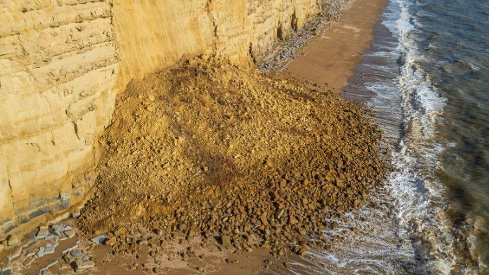 Golden cliff face with rocks scatters below forming a large pile and waves from sea lapping at the edge of the fall to the right