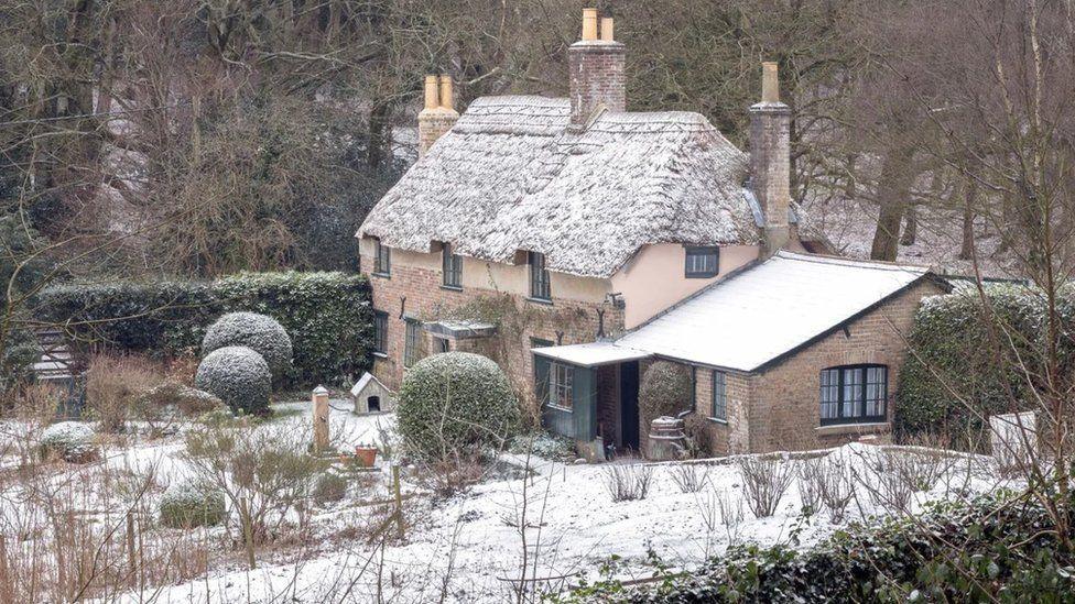 Thatched cottage standing in gardens with woodland behind - a sprinkling of snow can bee seen on the ground and the roof of the cottage.