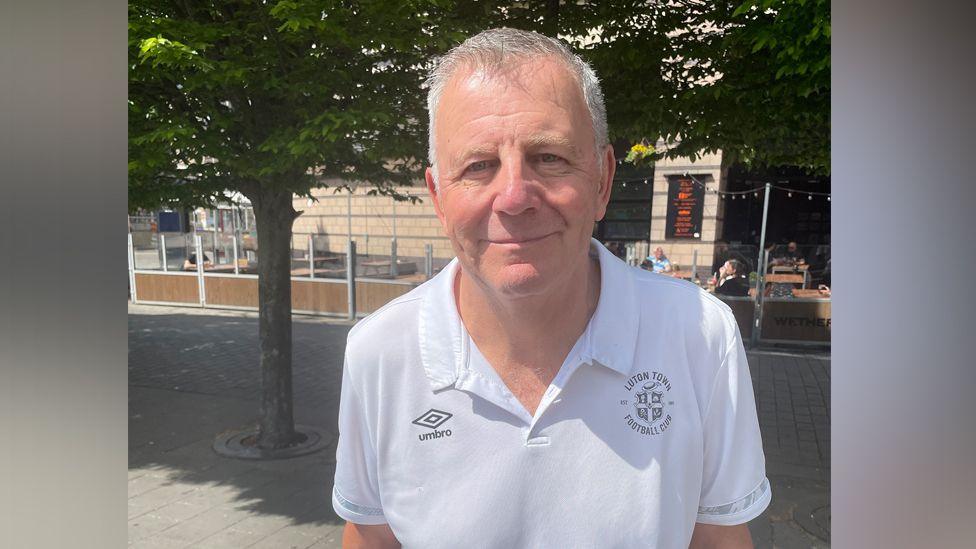 Paul Stephens standing in St George's Square in Luton, wearing a white collared t-shirt with the Luton Town logo on the left and Umbro logo on the right. He has grey cropped hair and smiles at the camera. There are trees behind him and outdoor cafe seating.
