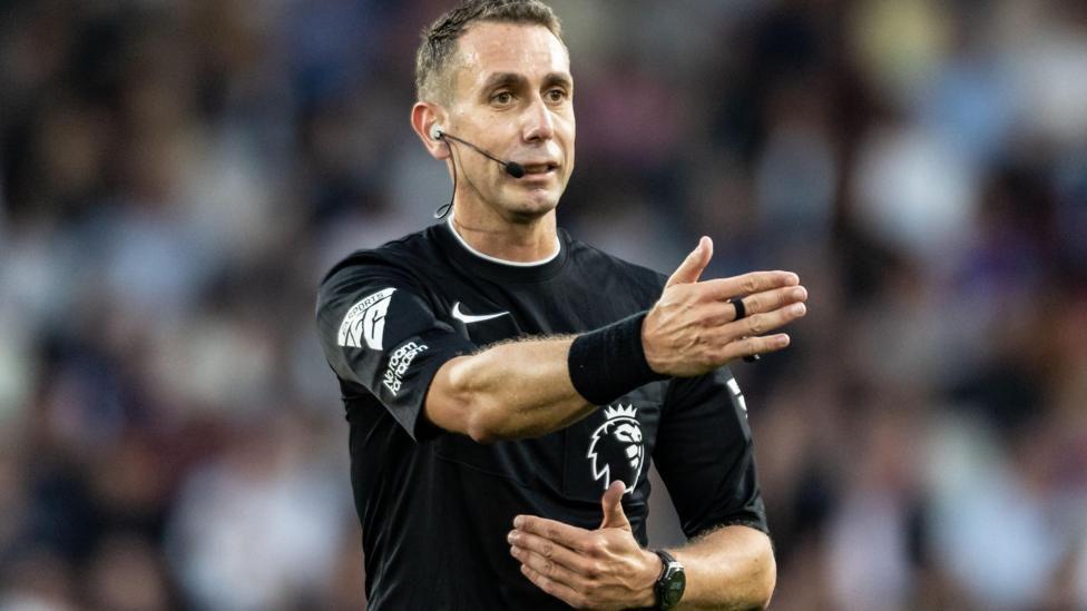 David Coote refereeing a premier league match. He is wearing and black football shirt and is holing one hand out in front of him as he makes a call in the game.