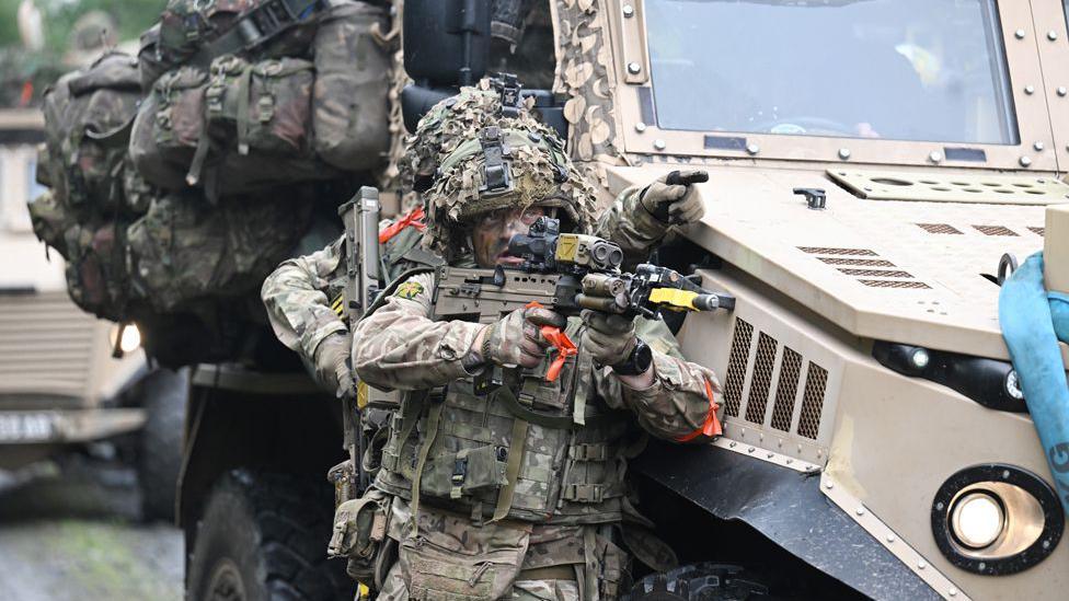 A solider aiming his rifle to the right of shot. His colleague behind him is pointing towards a target and they are leaning against an armoured vehicle.