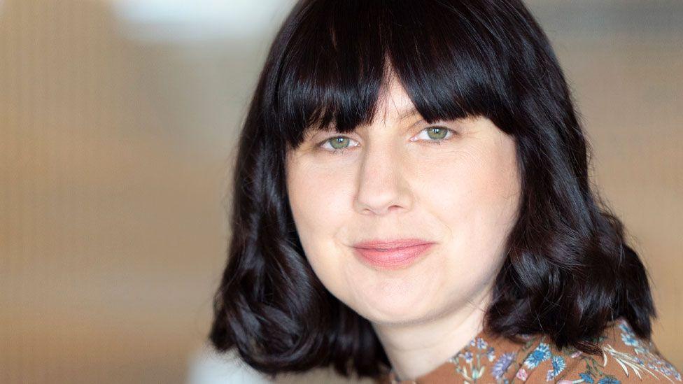 Dr Ceri Houlbrook, who has shoulder-length black hair with a long fringe. She is wearing a just-glimpsed brown top with blue and white flowers and leaves and is looking over her shoulder with the beginnings of a smile 