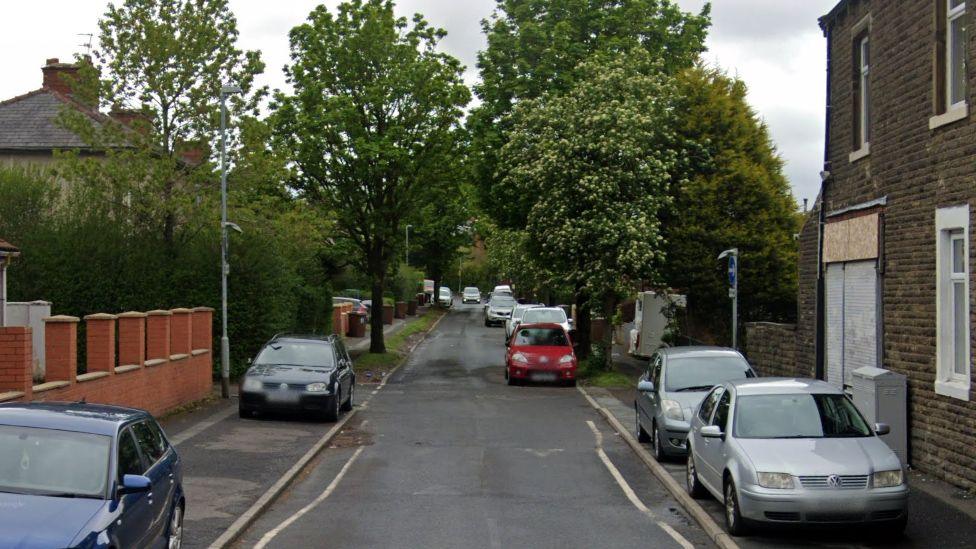 A street view image of Wordsworth Road with  terraced house to the right, a red brick wall to the left and trees in the distance, cars are parked on either side