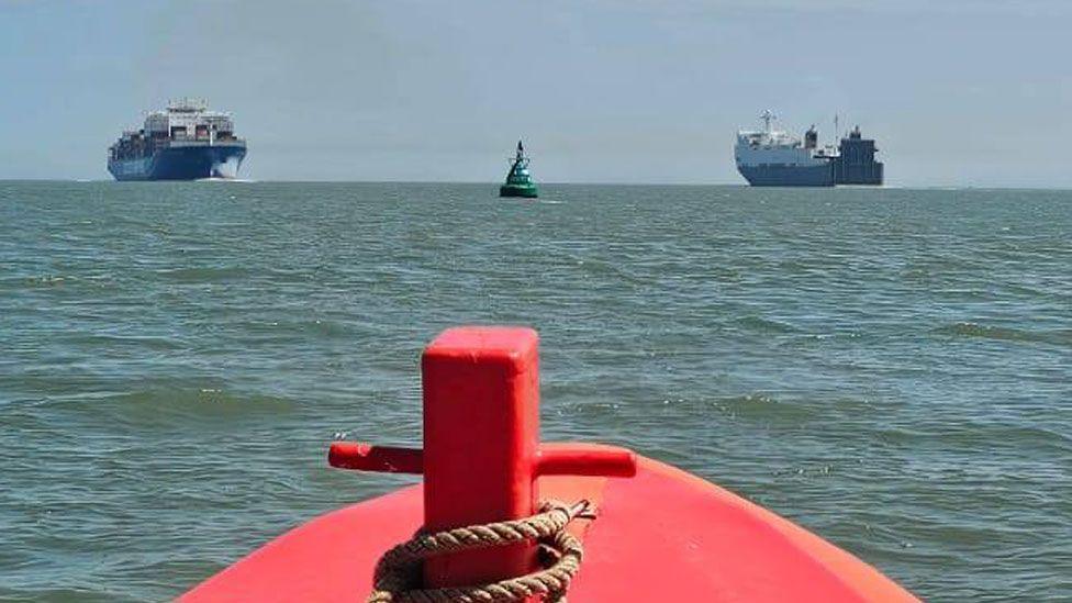 The red prow of a boat on greeny-blue seawater in the Thames Estuary. On the horizon are two huge container ships. In the middle is a large green buoy.