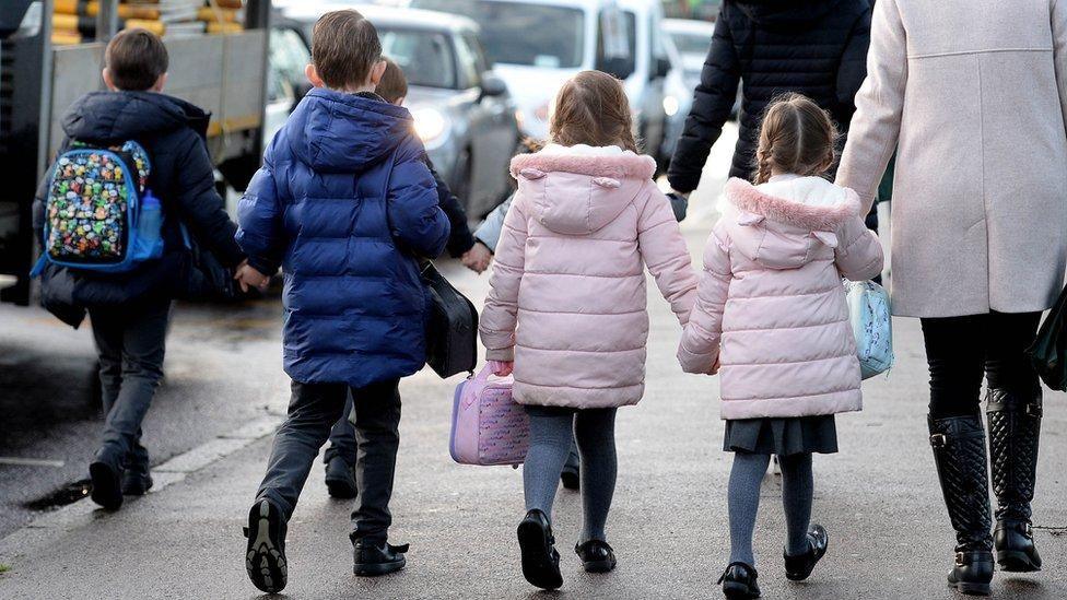 Five primary school age children walking on a street. There are three boys, wearing blue jackets, and two girls wearing pink jackets. The girls are holding hands, and one of the girls is holding hands with an adult who is also wearing a pink jacket.