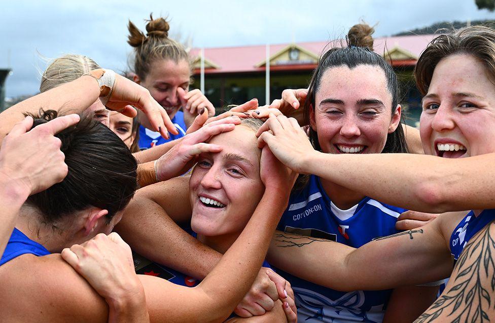 A group of female Australian rules footballers wearing blue sports jerseys huddle closely together, arms extended inward with hands stacked in a gesture of unity after a game. The background shows a building with a red roof and cloudy sky.