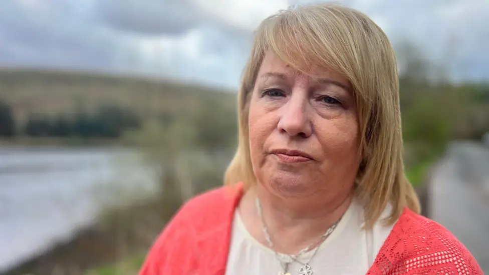 Maxine Johnson has blonde hair. She is looking directly into the camera, and is wearing a white t shirt and a pink cardigan