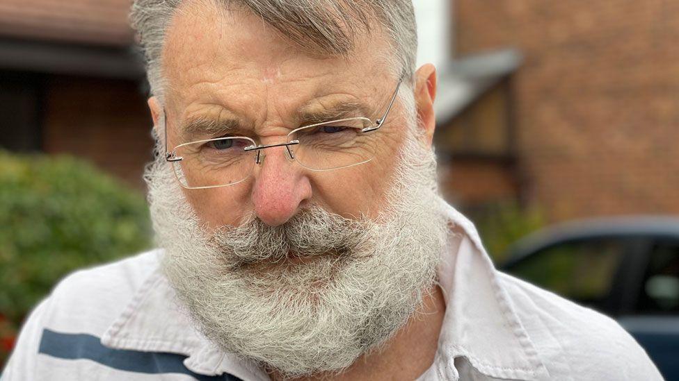Stephen Battle has grey hair, a white beard and is wearing rimless glasses. He is looking directly at the camera. He is wearing a white shirt with a blue stripe. Behind him are red brick houses and a parked car can just be glimpsed.