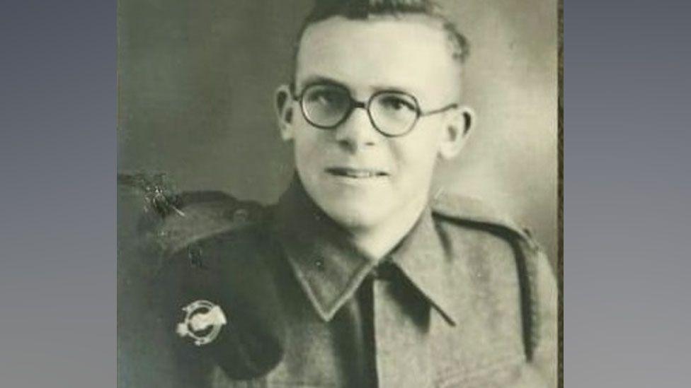 A black and white photo of Roy Hayward wearing World War Two Army uniform and round glasses, with his beret tucked up on his right shoulder, his head turned towards the camera 