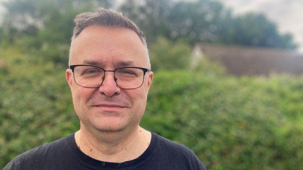 A man looking at the camera. He is wearing glasses and black T-shirt. A hedge is behind him.