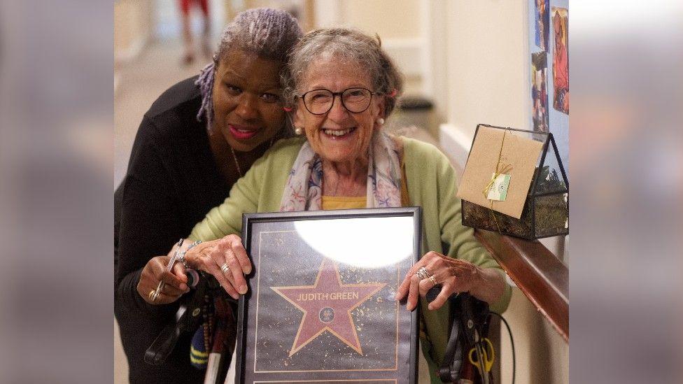 An elderly woman in a wheelchair smiles as she poses with a framed Hollywood Walk of Fame star with her name, Judith Green, on it. A lady is standing behind her wheelchair, with her arms around her.