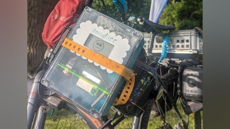 A clear plastic device with a screen and electric circuitry, with the word SmogOff and a smiley face on the screen. The device is attached to a bicycle.