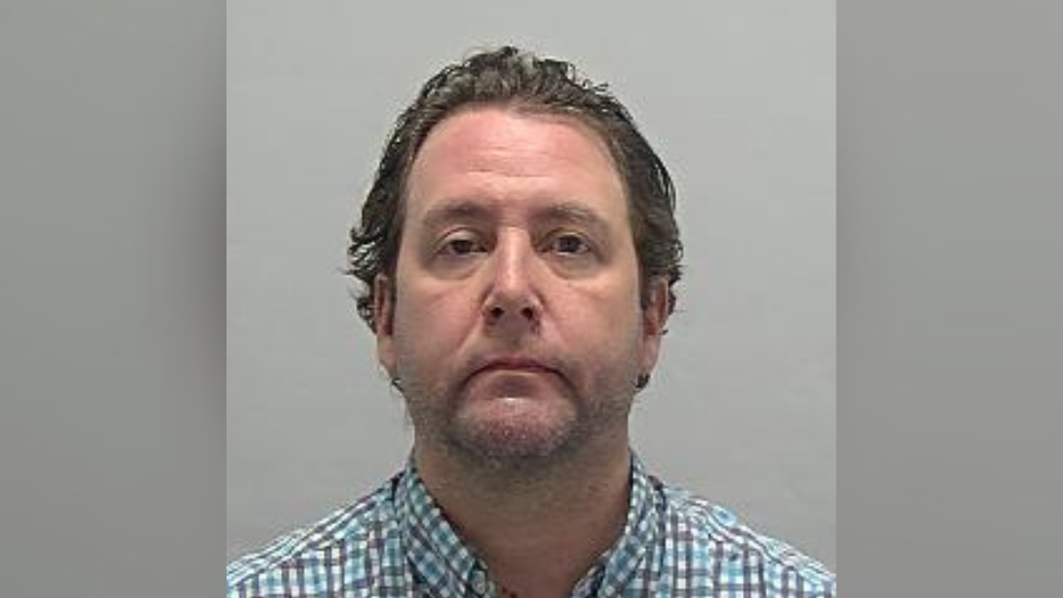 Custody photo of Patterson wearing a blue, white and grey checked shirt. He is stood against a grey background, has slight stubble on his face and dark brown hair. 