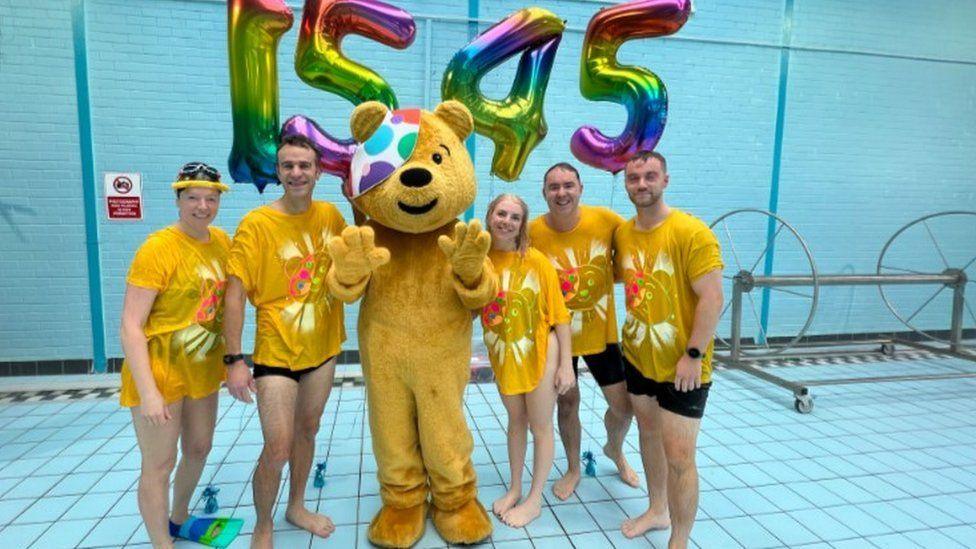 Numbered balloons, 1545, behind Pudsey bear - centre and BBC South and Radio Solent swimmers either side standing by a swimming pool.