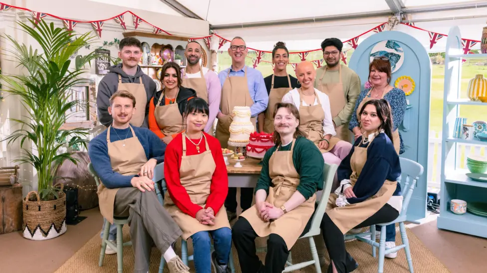 all the bake off contestants wearing aprons, sitting on chairs looking at the camera in the bake off tent