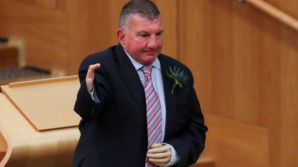 A man with short grey hair stands in the Scottish Parliament chamber. He is wearing a dark suit, blue chequered shirt and pink stripy tie. He has a prosthetic left hand.