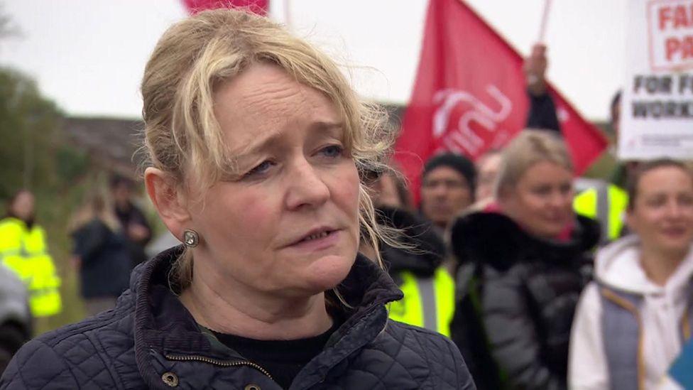 Unite general secretary Sharon Graham wearing a dark blue padded jacket stands in front of striking workers holding flags and banners