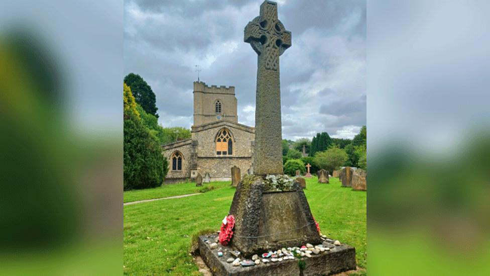 War memorial, with pebbles and a poppy wreath at its base, surrounded by lawn, with church and gravestones in background