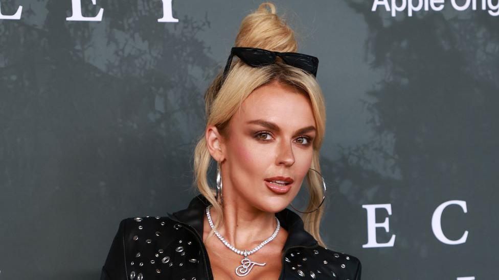Tallia is looking at the camera. She has blonde hair which is tied up. she has black sunglasses on the top of her head. She is wearing a sparkly necklace and a black top. and earrings.