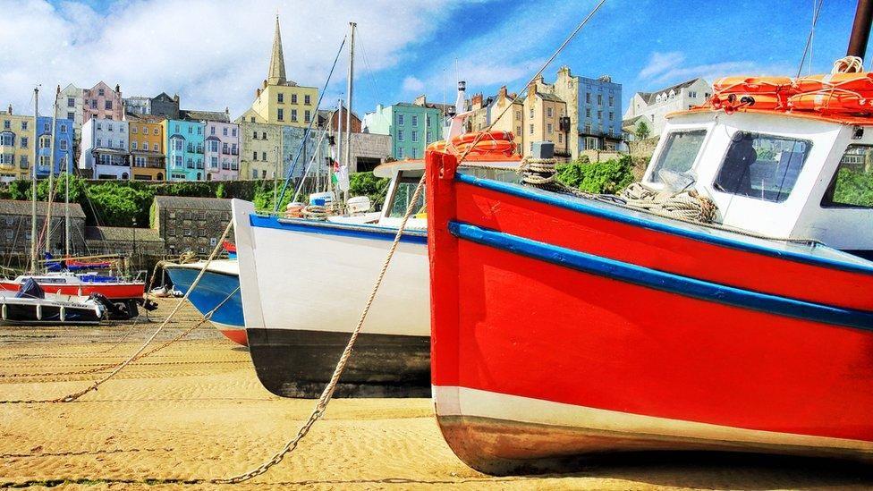 Picture of multicoloured houses in Tenby, overlooking the harbour at low tide. Several boats can be seen moored on the sand.