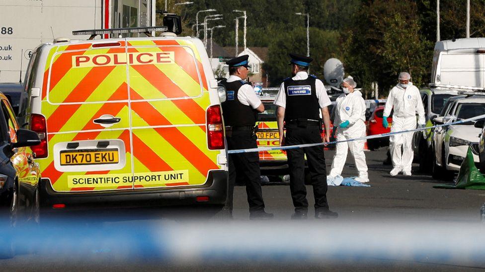 Two police officers stand next to each other beside a police van as forensics officers dressed in white overalls work on a street