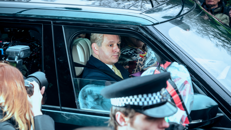 Michael Sheen as Prince Andrew. He is in a car and there is a police officer and a photographer outside next to the vehicle.