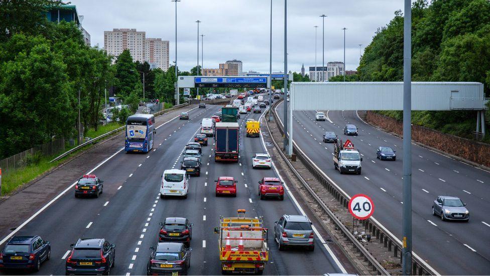 A general view of traffic on the M8 motorway, which has 10 lanes, five in either direction. High rise buildings can be seen in the background, with leafy green trees bordering the road. 