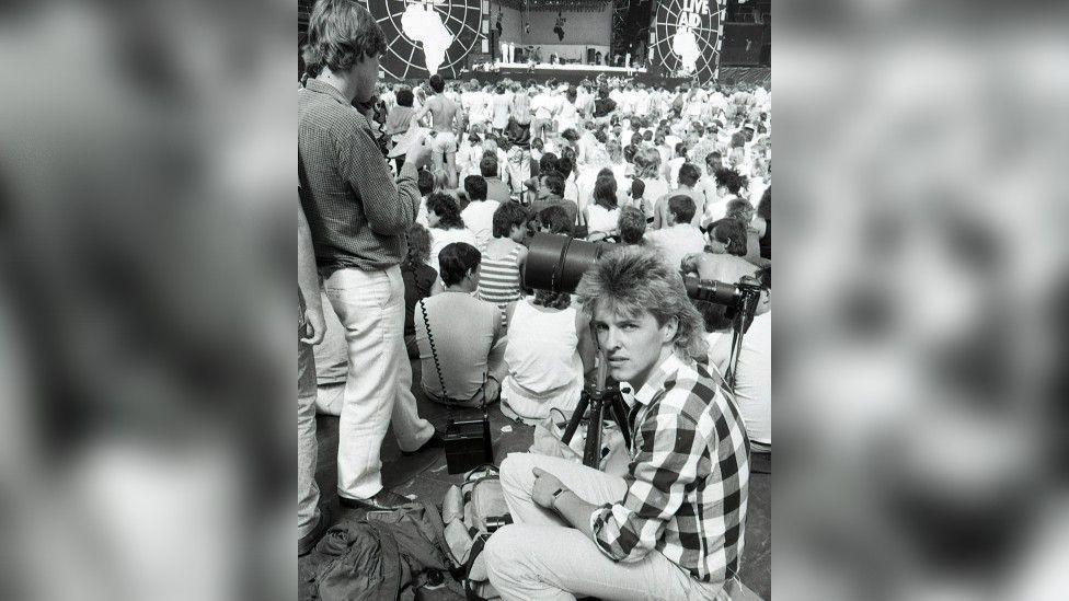 A black‑and‑white image  showing Murray Sanders wearing a checked shirt and sitting on the ground next to a camera mounted on a tripod. There's a crowd and the Live Aid stage behind him.