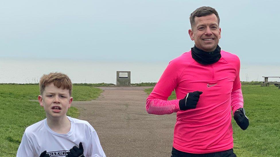 Ralphy and Stuart Raffle, who are running in Harwich. Ralphy is aged 10 and has short reddish hair and is wearing a white T-shirt. On his left is Stuart who has short grey hair and is wearing a pink running top and black gloves and neck tube/snood. Behind them is a wide path with grass on either side and the sea.