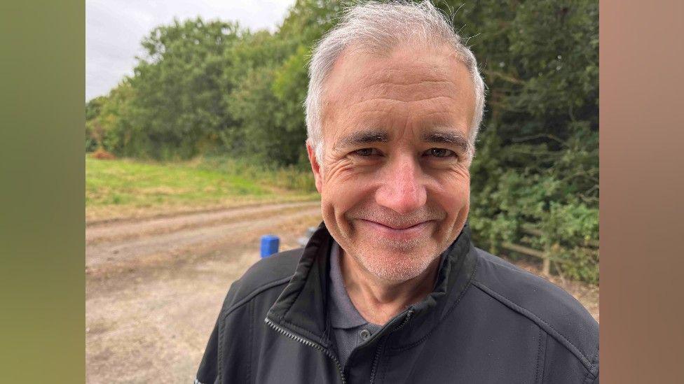 A man with grey-white hair is smiling at the camera. He has a black zip jacket on, and is standing on a gravel road with green grass and trees beyond